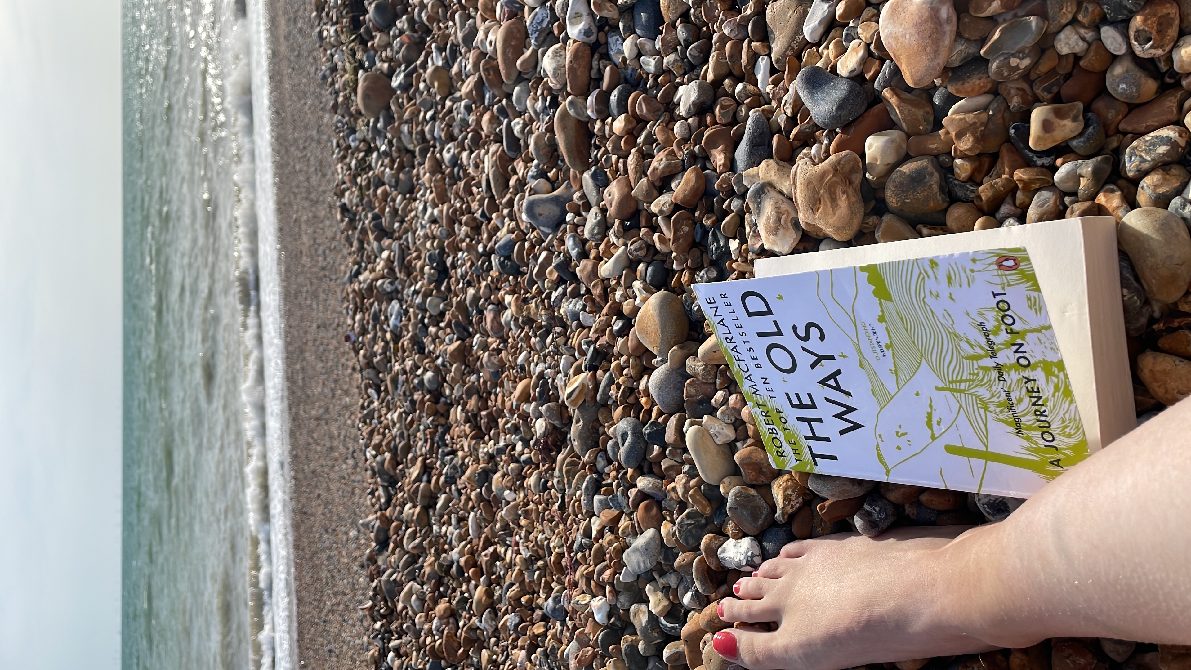 Image of a foot next to Robert Macfarlane's 'The Old Ways' on a pebble beach